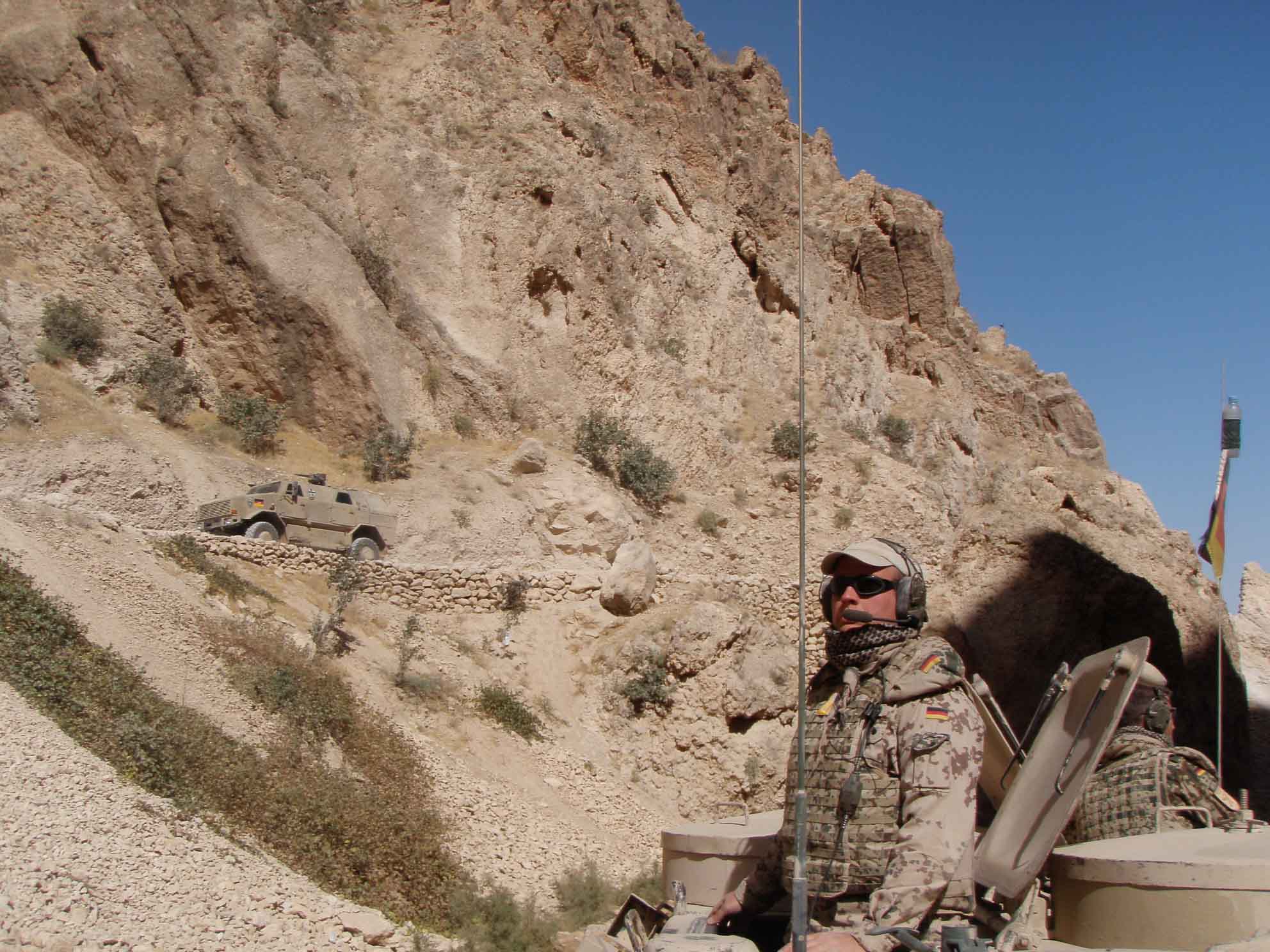 Ein Soldat in militärischer Ausrüstung sitzt in einem gepanzerten Fahrzeug in einer gebirgigen, trockenen Landschaft.
