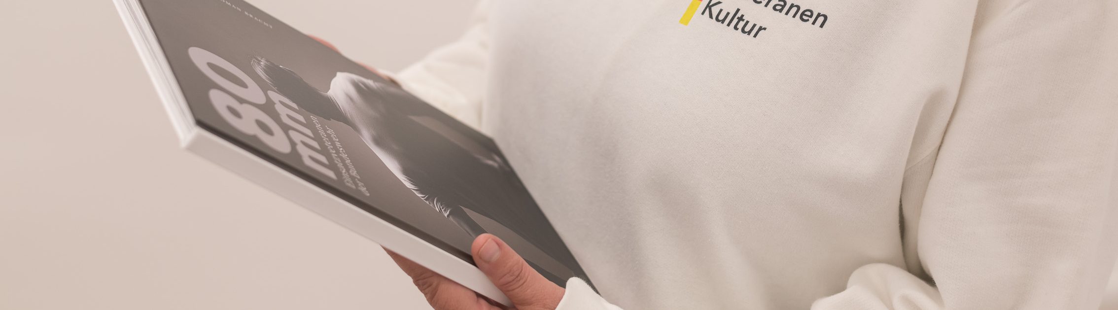 Eine Person hält ein Buch mit dem Titel "80 mm" und trägt ein Sweatshirt mit dem Logo "Kulturelle Bildung".