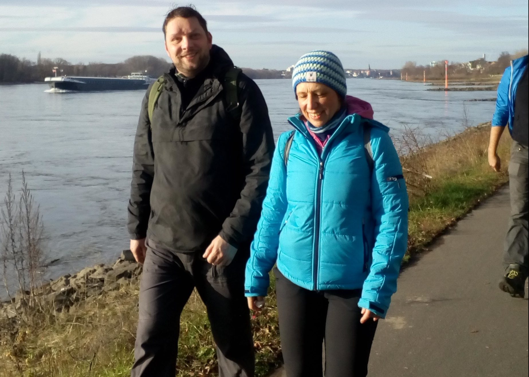 Zwei Personen spazieren am Ufer eines Flusses entlang, umgeben von Natur und klarer Winterluft.