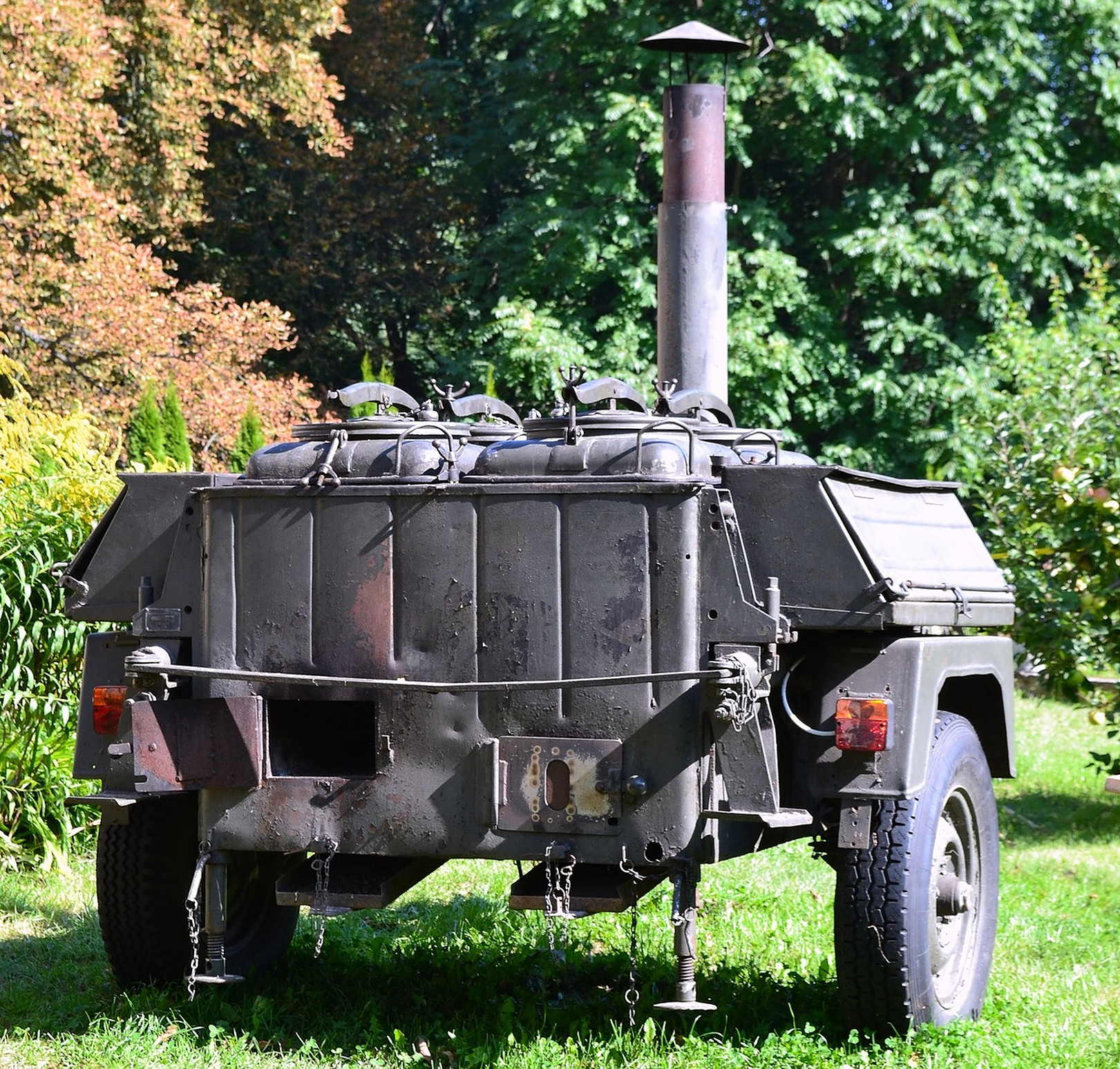 Ein schwarzer Anhänger mit einem Schornstein und mehreren Behältern, umgeben von grüner Vegetation.