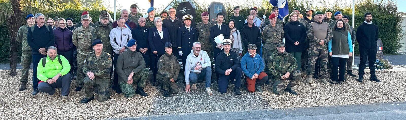 Gruppenfoto von Veteranen und Angehörigen in Uniform und Zivilkleidung, versammelt an einem Denkmal im Freien.